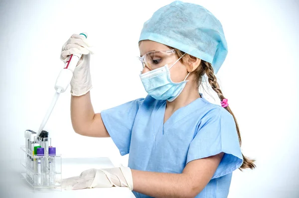 En liten flicka läkare i en medicinsk cap, mask, plast skyddsglasögon och handskar håller en dispenser i händerna och drar vätska från en medicinsk provrör. — Stockfoto