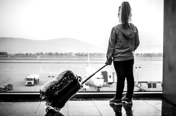 A black-and-white photo of a little girl standing with a suitcase at the airport and looking at her plane.