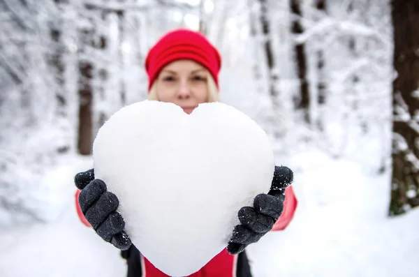 The girl is holding a heart from the snow. Heart in focus against the background of a blurry girl.
