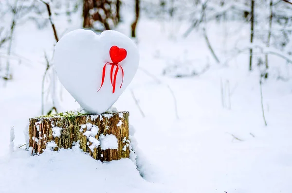 A big heart of snow stands on a stump. It has a small red heart with ribbon.