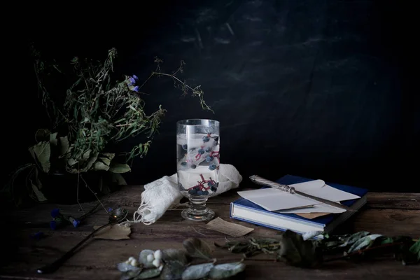 Stilleven, vintage. een glas mineraalwater en bessen, boeken, boeket op houten tafel. donkere achtergronden — Stockfoto