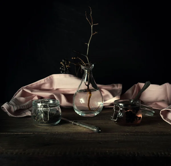 Vida morta rústica. galho seco em um vaso de vidro, cortinas rosa e jarros em uma mesa de madeira. fundo preto — Fotografia de Stock