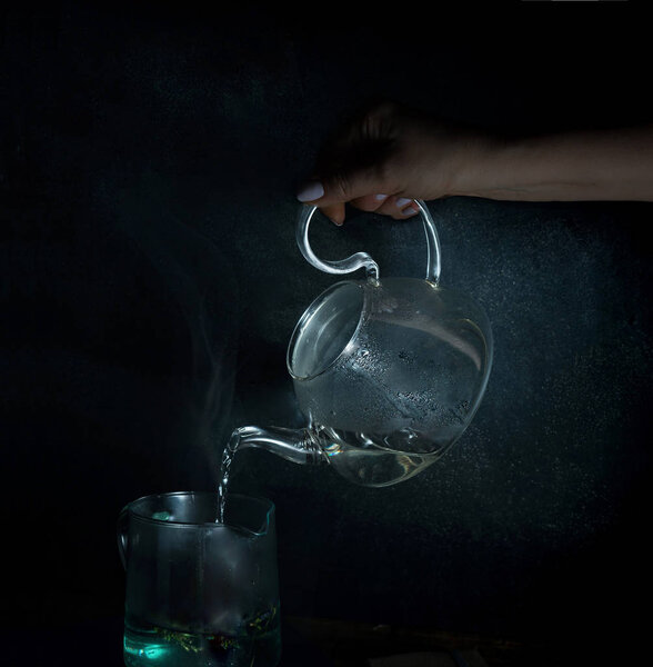 womans hand pours boiling water into a glass kettle. dark background. vintage.