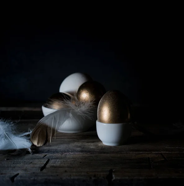 Pasqua. La notte di Pasqua. Uova d'oro e bianche, piume su un tavolo di legno. Vintage. Sfondo scuro — Foto Stock