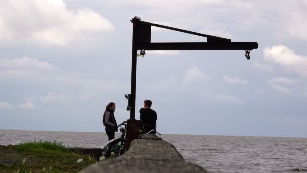 Los jóvenes y la chica se sientan en el muelle de piedra cerca de la bicicleta hablando sobre sus temas favoritos — Vídeo de stock