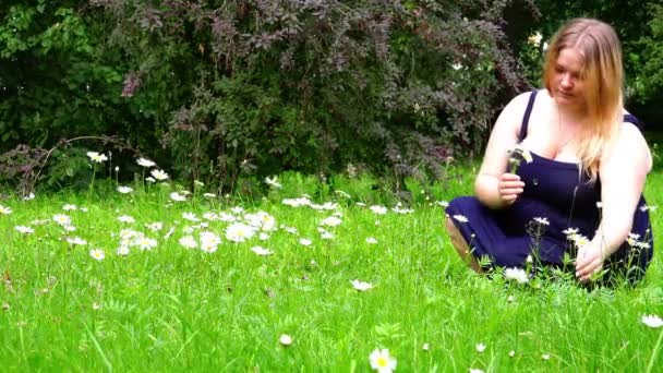 Una chica en un sarafán azul recoge flores silvestres sentadas sobre hierba verde — Vídeos de Stock