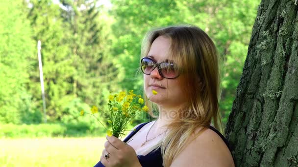 En flicka i solglasögon i frisk luft andas in doften av vilda blommor i parken — Stockvideo