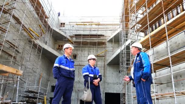 Werknemers in witte helmen en blauwe uniformen bespreken werken momenten tegen een monolithische structuur van gewapend beton — Stockvideo