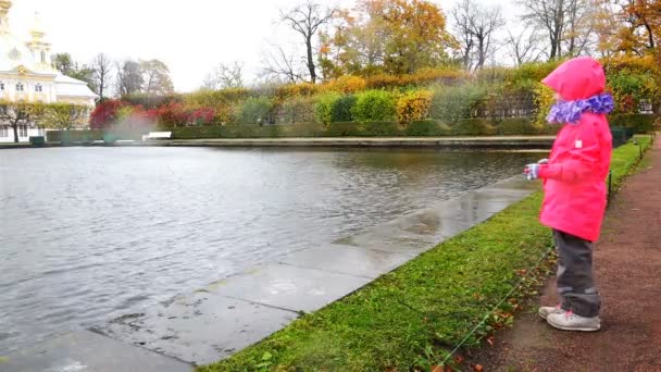 Een meisje in een rode jas is permanent in de regen in de buurt van de oever van een vijver in het park-zone — Stockvideo