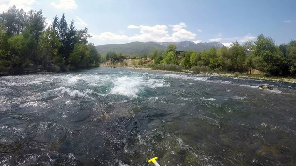 Bir adam botun ön tarafında oturuyor. Bacakları sallanıyor ve bir dağ nehrinde rafting yapıyor.