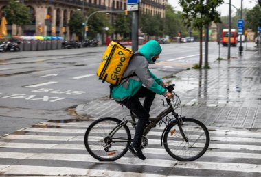 Barselona, İspanya. 20 Nisan, 2020. Glovo teslimatçısı yağmur altında Barcelona 'nın boş sokaklarında bisiklet sürüyor.