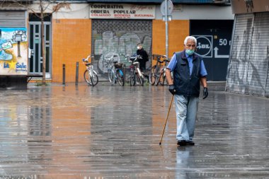 Barselona, İspanya. 20 Nisan, 2020. Koruyucu yüz maskesi takan yaşlı bir adam ve tamamen boş bir Barceloneta meydanında yürüyorlar.