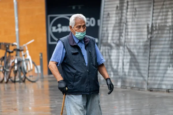 Barselona, İspanya. 20 Nisan, 2020. Koruyucu yüz maskesi takan yaşlı bir adam ve tamamen boş bir Barceloneta meydanında yürüyorlar.
