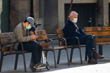 Barselona, İspanya. 4 Mayıs 2020. Orta Barselona 'da yaşlı bir adamın yanındaki bankta oturan genç adam yaşlıların dışarı çıkmasına izin veriliyor.