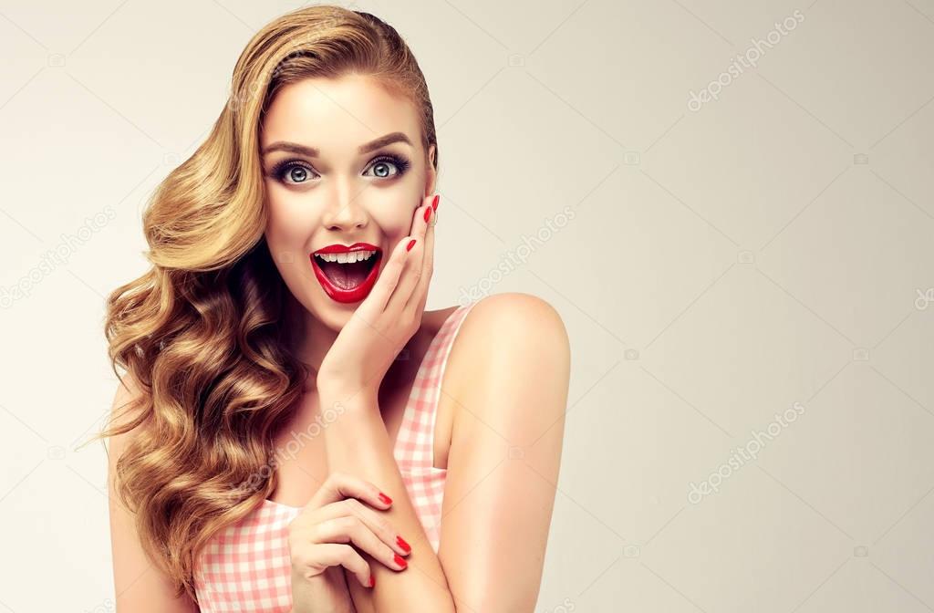 Woman with red lips and nails surprise holds cheeks by hand .Beautiful girl  with curly hair surprised and shocked looks on you . Presenting your product. Expressive facial expressions