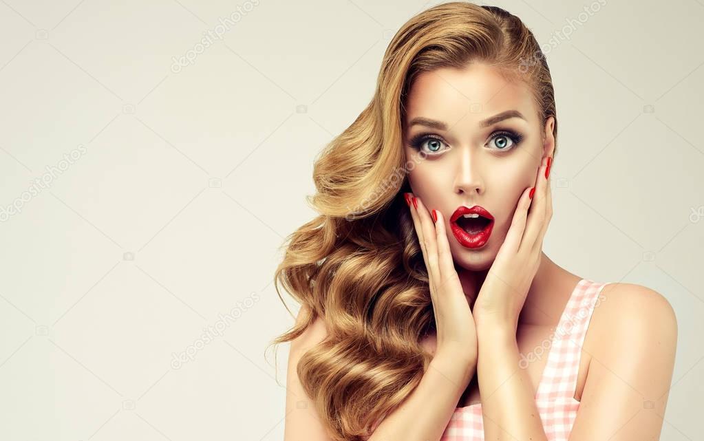 Woman with red lips and nails surprise holds cheeks by hand .Beautiful girl  with curly hair surprised and shocked looks on you . Presenting your product. Expressive facial expressions