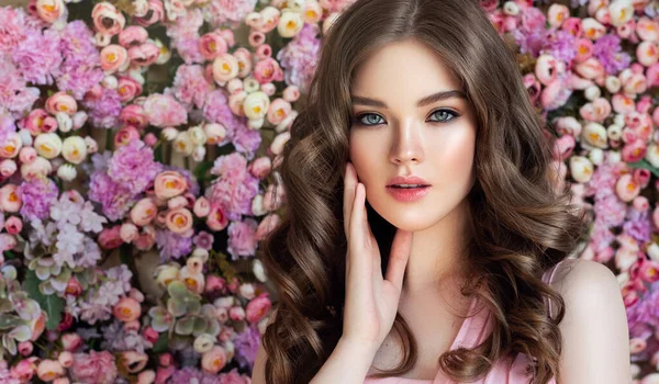 長く光沢のある波状の髪を持つブルネットの春の女の子 巻き毛の美しいモデルの女性 花の背景の壁 スキンケア — ストック写真