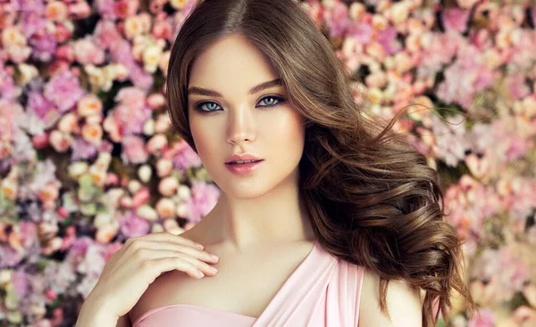 長く光沢のある波状の髪を持つブルネットの春の女の子 巻き毛の美しいモデルの女性 花の背景の壁 スキンケア — ストック写真