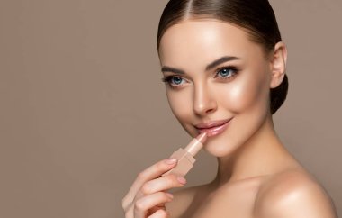 Beautiful young woman smears a balm on her lips. The girl uses hygienic nude lipstick. Cosmetics, makeup and skin care. clipart