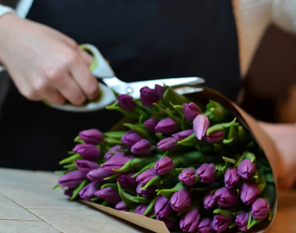 Una mujer hace un ramo de flores en la mesa. Tulipanes lila, envueltos en papel . — Foto de Stock