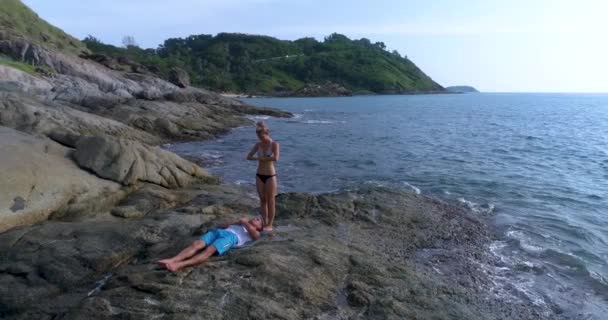 Aérienne : Un beau jeune couple pratique le yoga sur les rochers à côté de la mer — Video