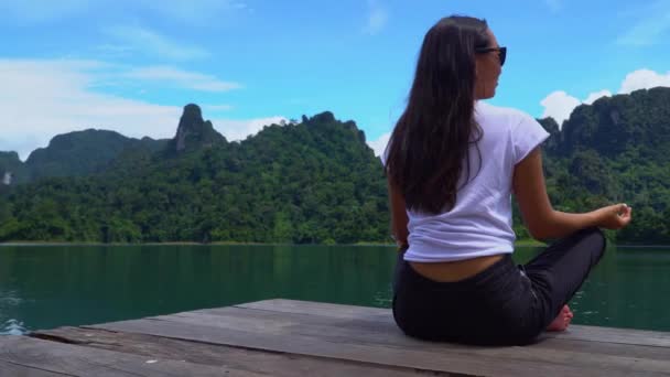 Asiática chica se sienta en un muelle en una pose de loto en un lago con una hermosa vista . — Vídeos de Stock