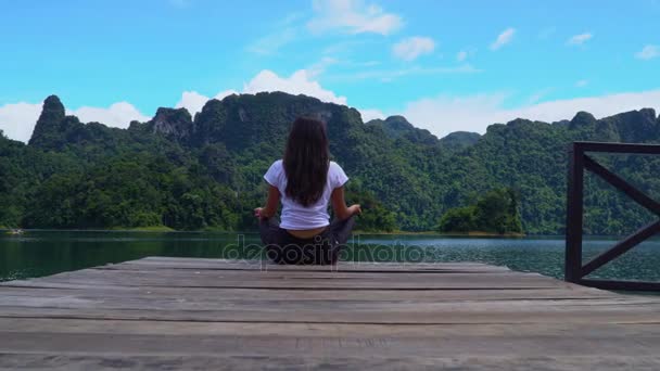 Uma menina senta-se em um cais em uma pose de lótus em um lago com uma bela vista . — Vídeo de Stock