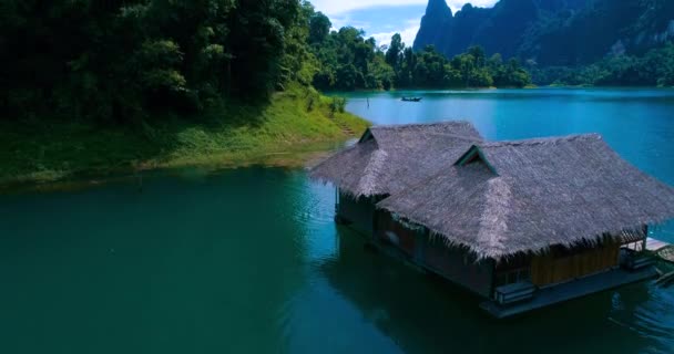 Aéreo: Uma casa com um telhado de palha no lago e um barco navegando ao lado . — Vídeo de Stock