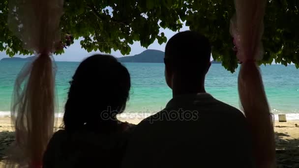 Νεαρό ζευγάρι κάθεται σε ένα σχοινί ταλάντευσης στην παραλία κάτω από τα δέντρα. — Αρχείο Βίντεο
