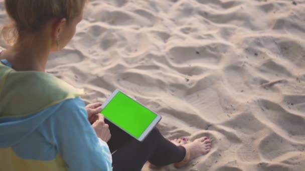 Een meisje zit op het zand en het gebruik van een tablet met chroma key scherm. Jatten. — Stockvideo