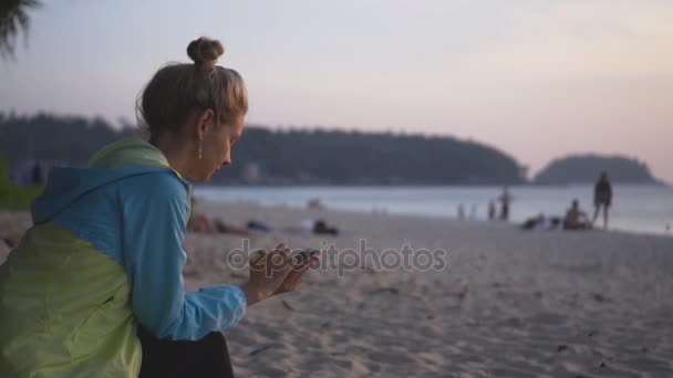 Menina triste com um telefone senta-se na praia ao pôr do sol . — Vídeo de Stock