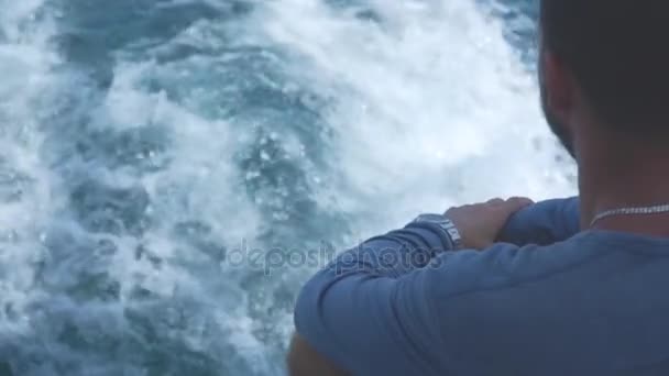 Young man sails on a yacht in a not calm sea and looks at a ship on the horizon. — Stock Video