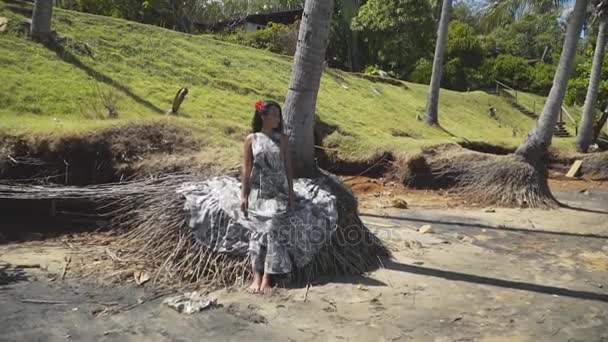 Gri elbiseli bir palmiye ağacının kökleri üzerinde oturan ve poz Asyalı kız. — Stok video