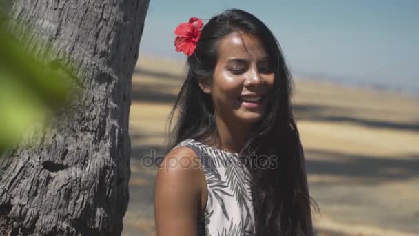 Peeping after an Asian girl with red flower in hair laughing at the beach. — Stock Video