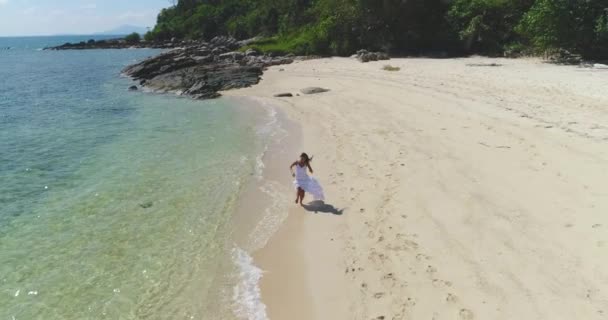 Aéreo: Uma menina em um vestido branco está correndo ao longo da praia pela água . — Vídeo de Stock