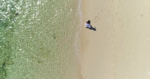 Aerial: Menina morena em um vestido branco está correndo ao longo da praia pela água . — Vídeo de Stock