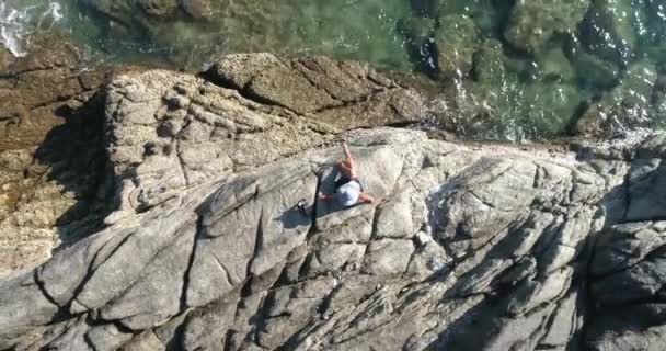 Aéreo: Uma mulher de chapéu senta-se na praia em rochas e olha para o mar . — Vídeo de Stock