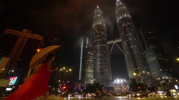Kuala Lumpur, Maleisië 13 november 2019: Een meisje in een rode jurk met een paraplu loopt 's nachts in de buurt van de Petronas Towers. — Stockvideo