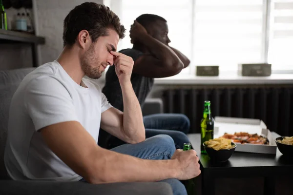 Disappointed guy watching football match with African American friend
