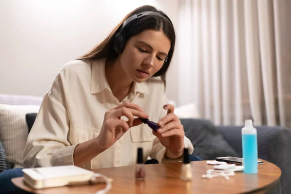 Young female listening to music in headphones and closing bottle with dark blue nail polish while siting at table with smartphone and manicure supplies at home