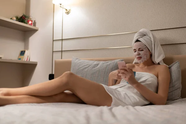 Young female in skincare mask and wet towels enjoying hot beverage and surfing Internet on smartphone while resting on comfortable bed after shower in modern apartment