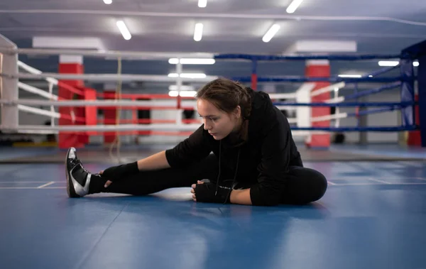 Full body strong woman in black sportswear sitting on floor and doing stretching exercise while preparing for sparring during boxing training in gym