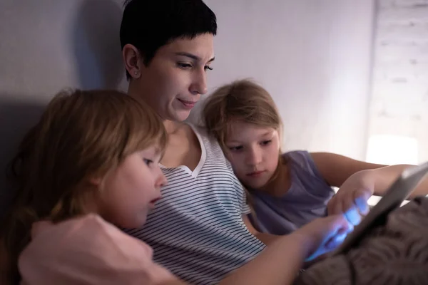 Little sisters playing games on digital tablet while lying on bed near mother before sleep in evening at home