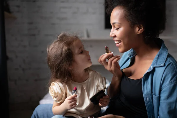 Delighted ethnic woman smiling and showing cute mixed race girl how to apply lipstick during makeup lesson at home