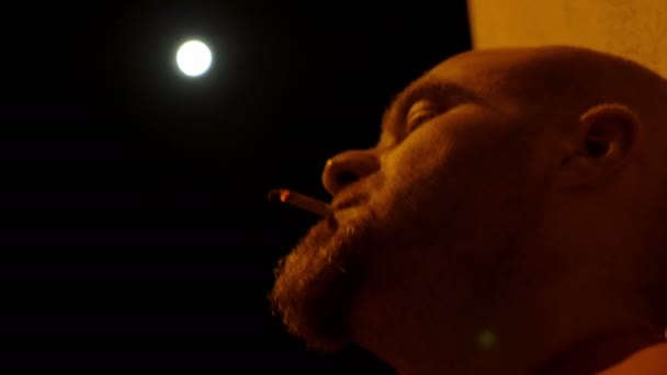 Bald man alone smoking a cigarette lit by the moon — Stock video