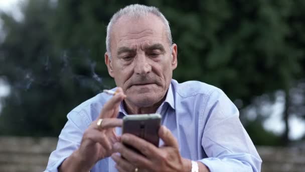 Elderly man at the park smoking a cigarette typing on mobile phone — Stockvideo