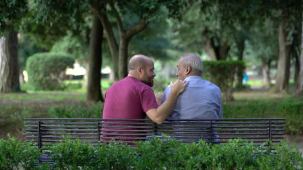 Padre e hijo sentados en un banco del parque hablando amablemente — Vídeos de Stock