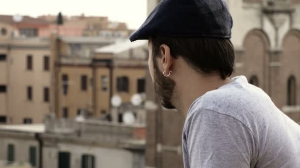Man met een oorbel en hoed vanaf het balkon met uitzicht op de stad — Stockvideo