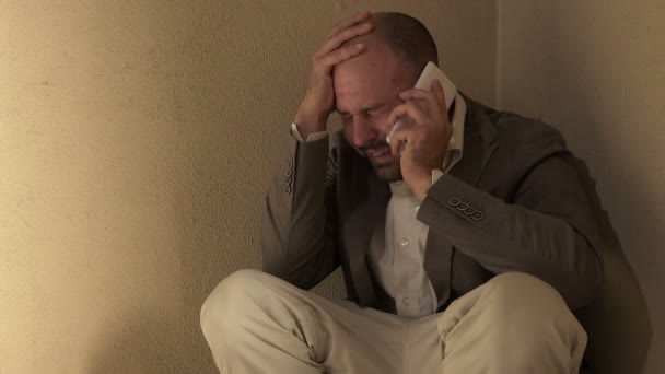 Человек отчаянно плачет разговаривая по телефону, приседая на полу — стоковое видео