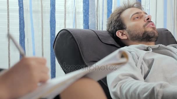 Hombre de 30 años acostado en el sofá hablando con su psicoanalista que toma notas — Vídeo de stock